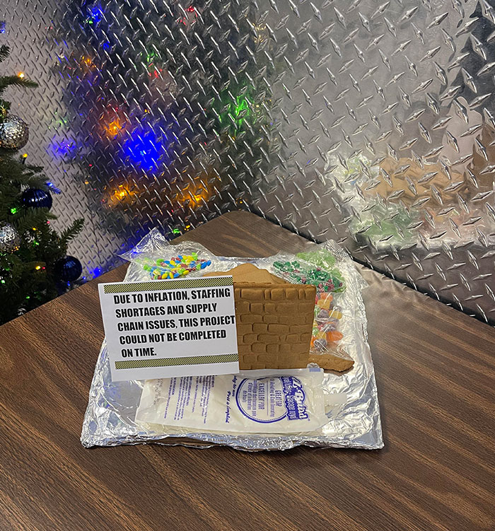 Christmas humor: Incomplete gingerbread house with a sign blaming inflation and staff shortages, festive lights in background.