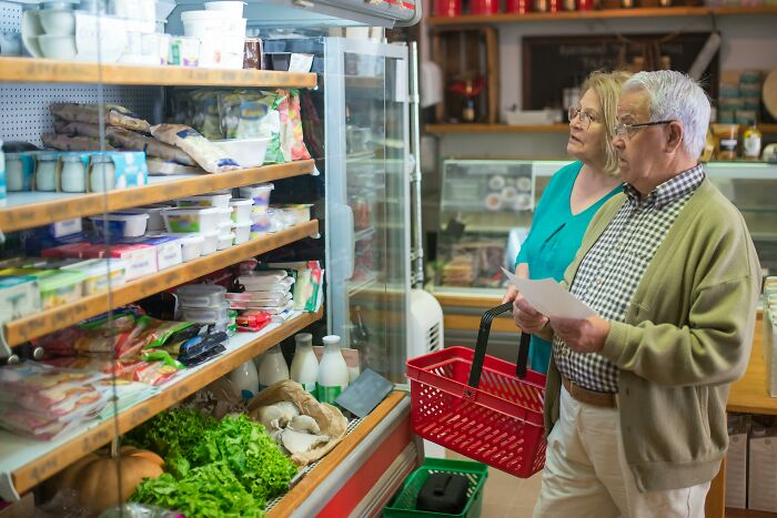 Elderly couple grocery shopping, possibly discussing funny overheard conversations, holding a list and basket in a store aisle.