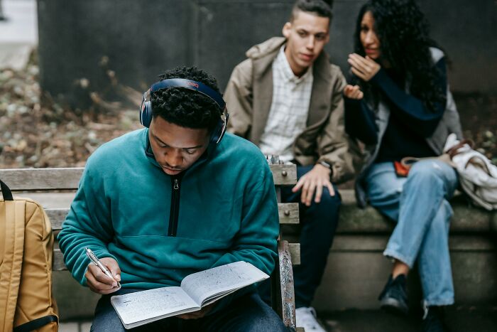 A person overhearing conversations on a bench, while writing in a notepad, others whisper behind.