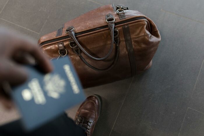Person holding passport near leather travel bag, possibly after hilarious overheard conversations at the airport.