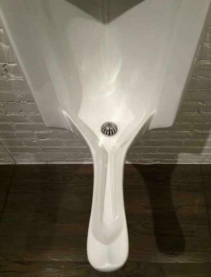 Unusual white urinal design that looks fake at first glance, set against a brick wall and wooden floor.