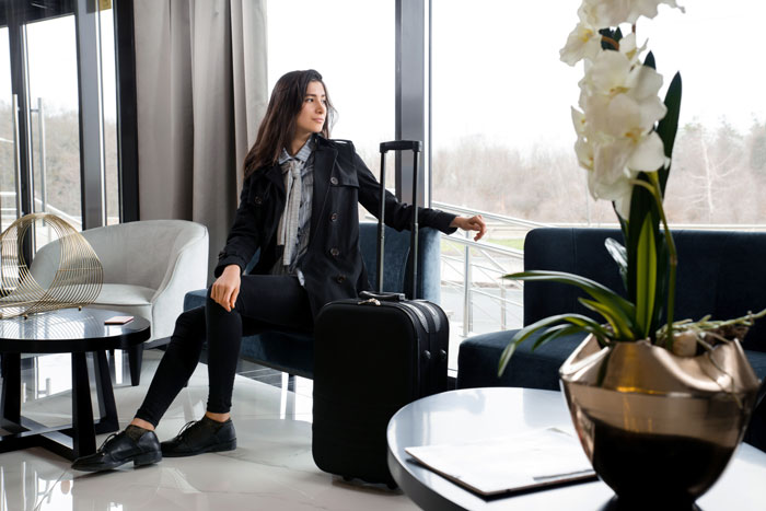 Young woman sitting in hotel lobby with suitcase, reflecting after sleep deprivation in relationship challenges.