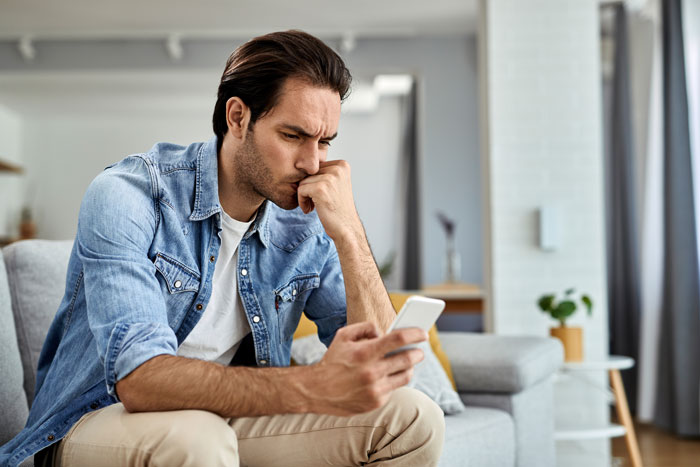 Man wearing denim shirt sitting on couch looking stressed while holding smartphone, illustrating sleep deprivation effects.