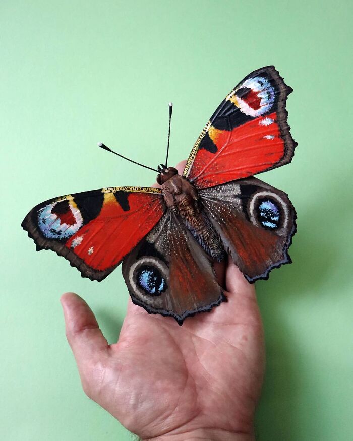Hand holding a lifelike butterfly crafted from paper and wood, showcasing detailed and colorful wings against a green background.