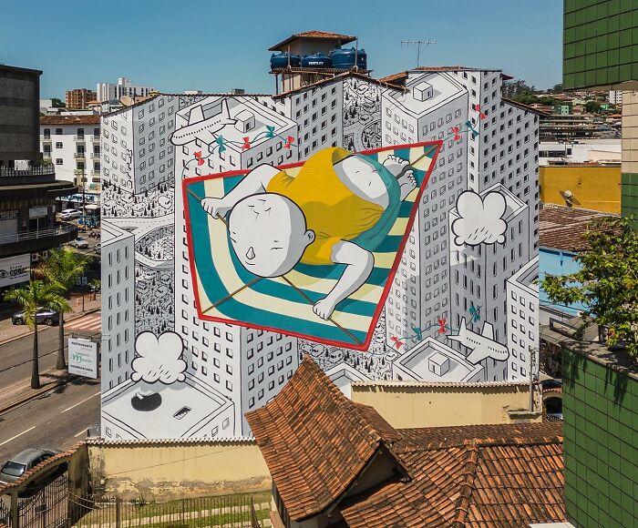 Colorful mural by Italian artist transforms city walls into whimsical world with a giant child flying over illustrated urban buildings.