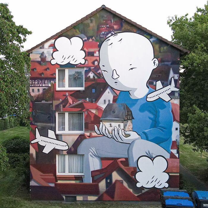 Mural by Italian artist depicting a large whimsical figure holding a house, surrounded by planes and clouds on a city wall.