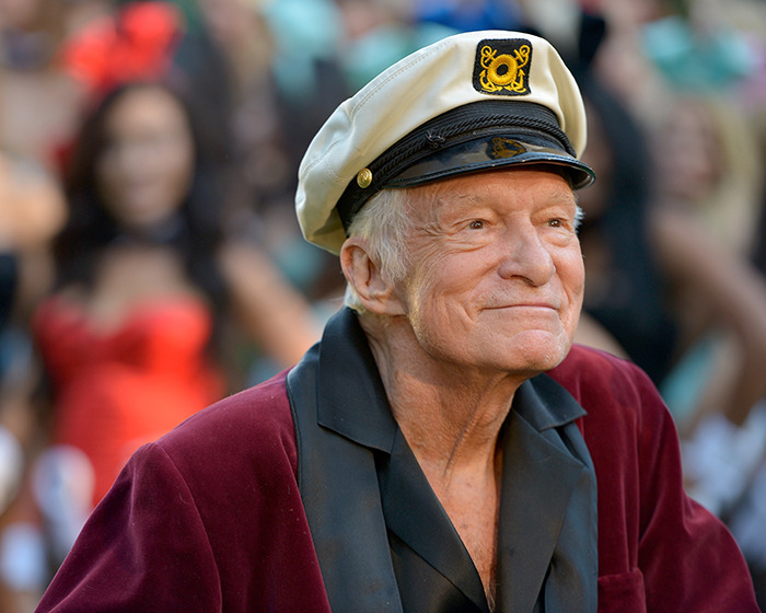 Hugh Hefner wearing a captain hat and velvet jacket, attending an outdoor event with blurred crowd in background.