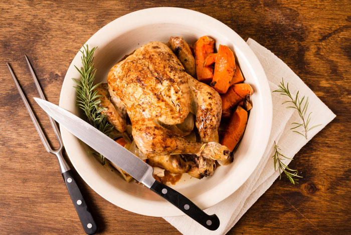 Roasted chicken with carrots on a white plate beside carving knife and fork on wooden table with herbs