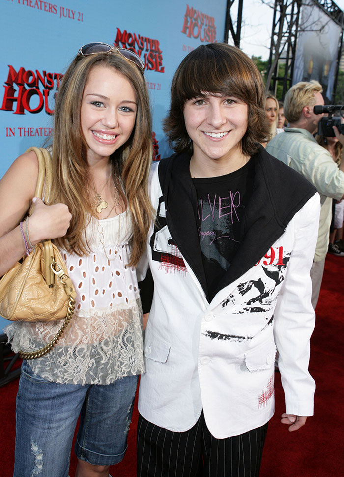 Miley Cyrus' sister Noah smiling in a revealing outfit at a red carpet event posing for photos.