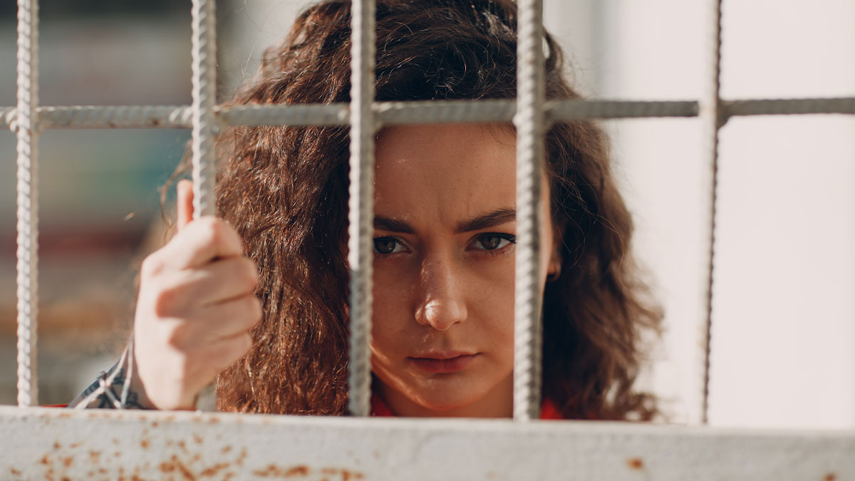 Young woman behind bars with serious expression, symbolizing crime and regret in a tense prison setting.
